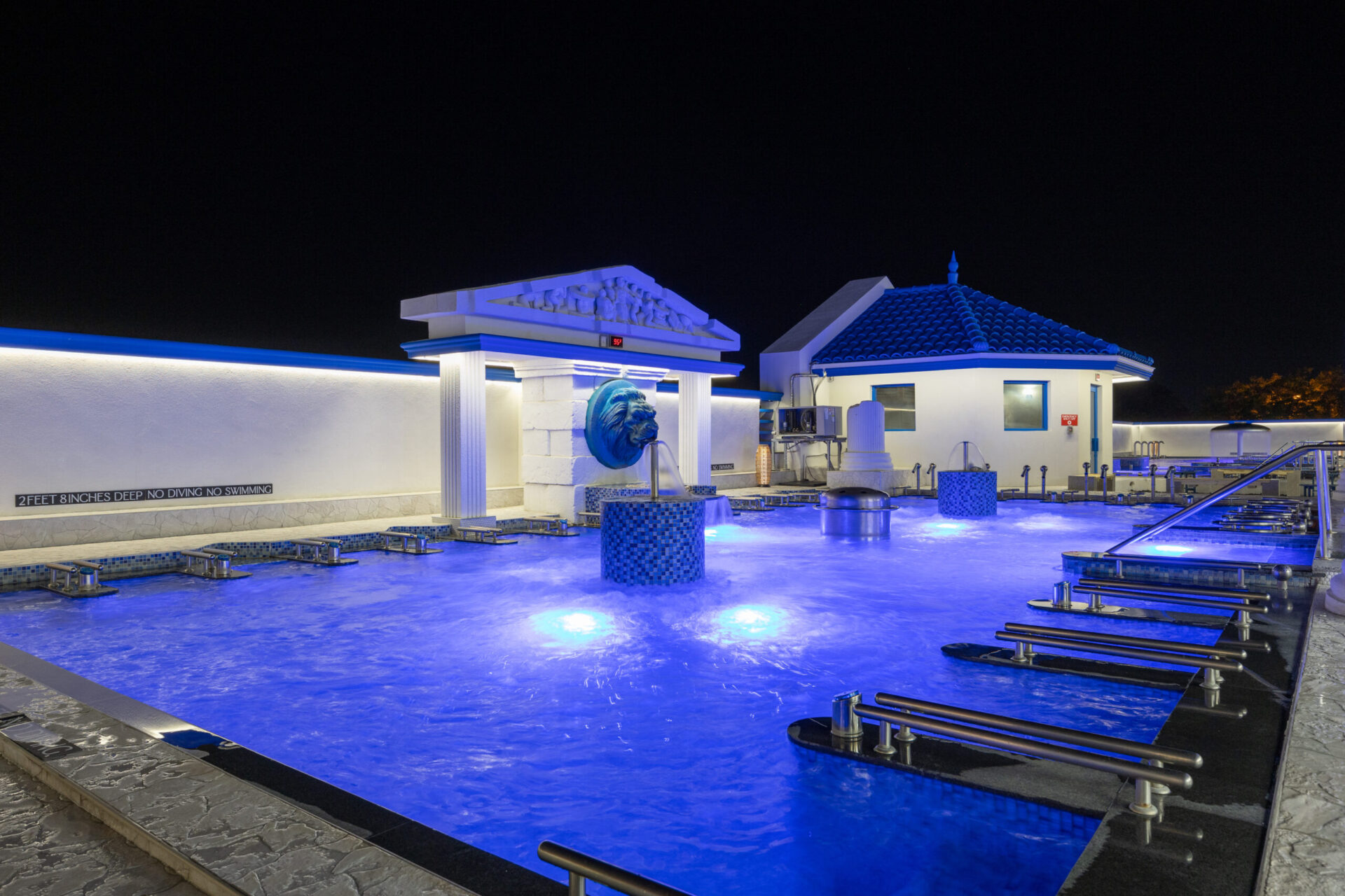 nighttime view of the outdoor seated medi pool. lit up with underwater lighting. With the seasonal sauna in view to the right