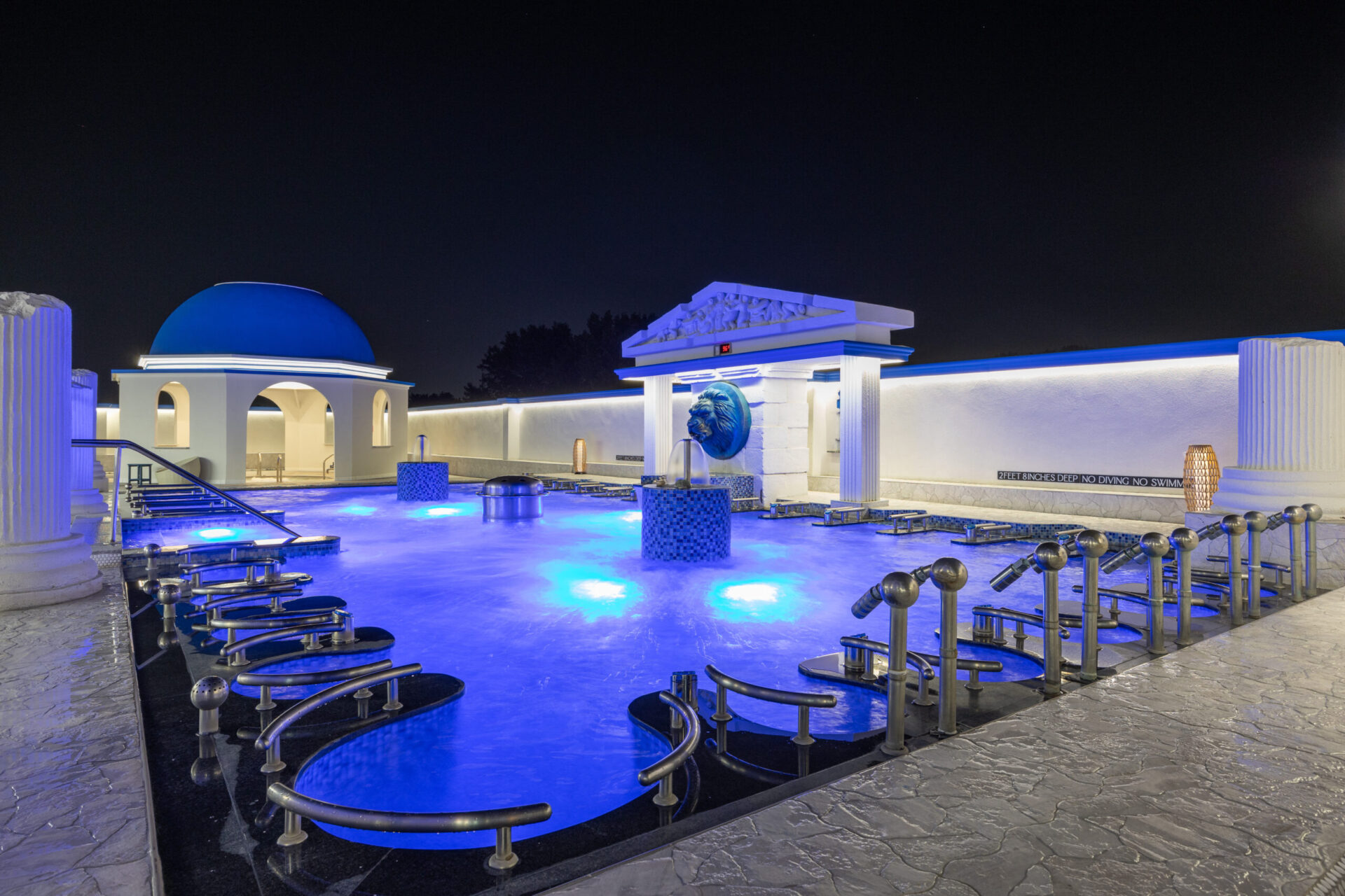 nighttime view of the outdoor seated medi pool. lit up with underwater lighting. With the Grotto Hot tub in view to the left.